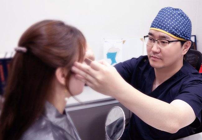 거울 앞에 앉은 여성 환자의 눈가를 살피는 원장(수술모·남색 수술복)